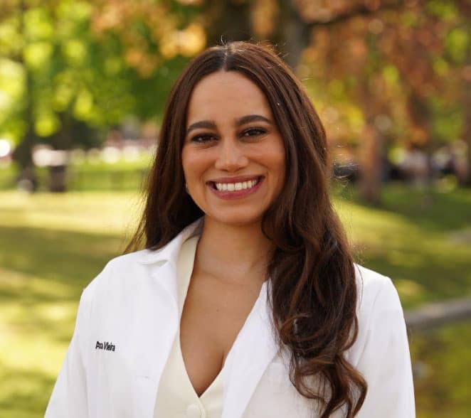 A woman with long brown hair, wearing a white lab coat, smiles while standing outdoors in a park with green trees in the background.